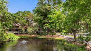 A serene pond with a fountain, surrounded by lush greenery and trees.