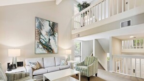 A spacious living area with a sofa, armchairs, a coffee table, and a large abstract painting. A staircase connects to an upper level with a balcony.