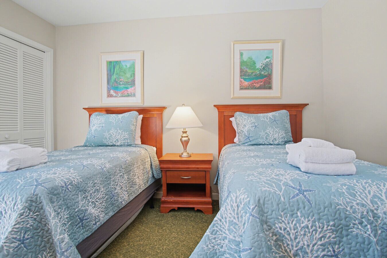 Habitación de hotel con dos camas, cada una con colcha azul adornada con motivos de estrellas de mar y coral, una mesita de noche de madera con lámpara, y dos cuadros enmarcados con paisajes.