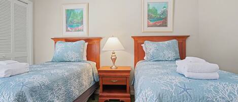 Habitación de hotel con dos camas, cada una con colcha azul adornada con motivos de estrellas de mar y coral, una mesita de noche de madera con lámpara, y dos cuadros enmarcados con paisajes.