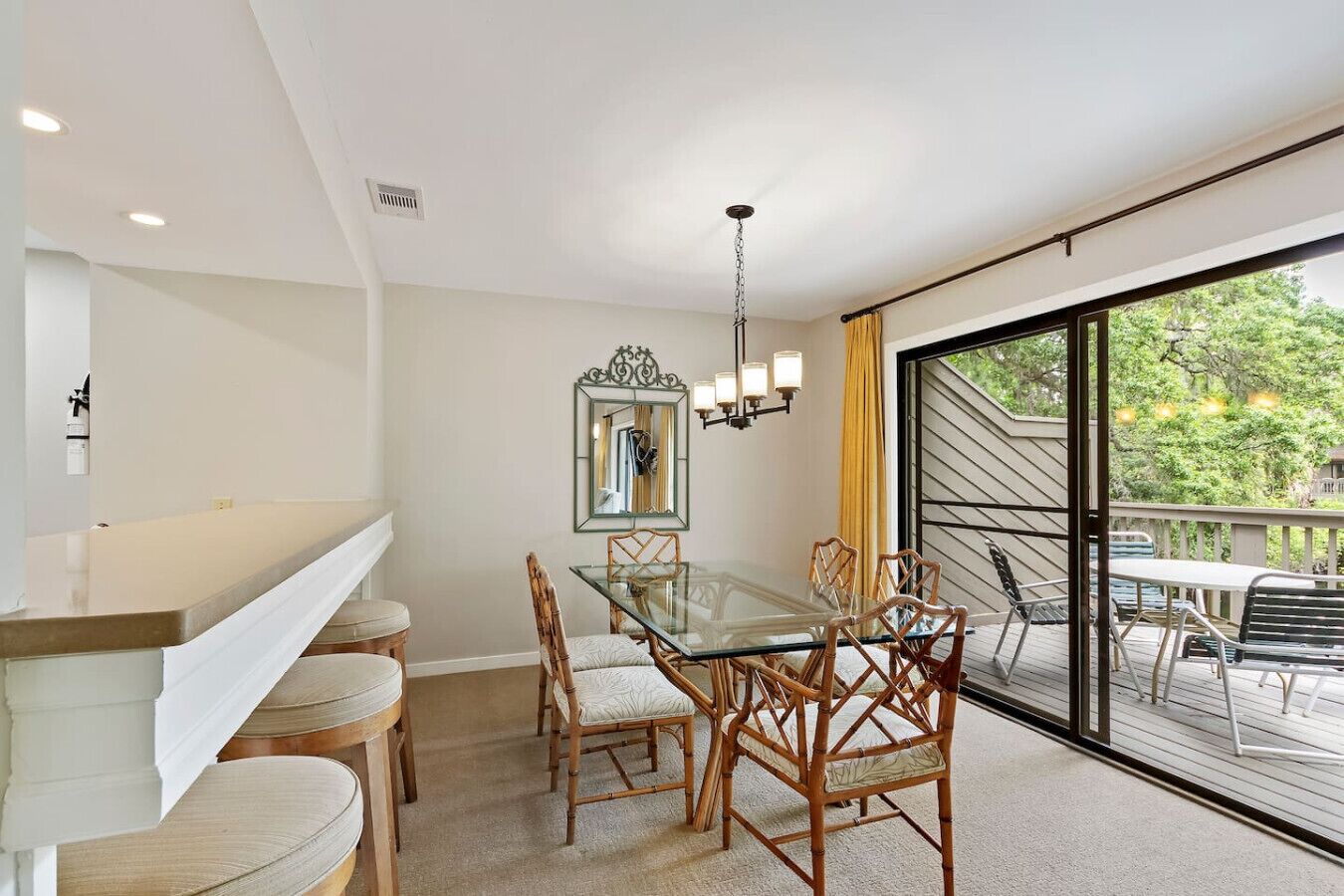 Un espace repas moderne avec une table en verre et des chaises en bambou, un grand miroir et une vue sur l’extérieur grâce à une porte-fenêtre coulissante.