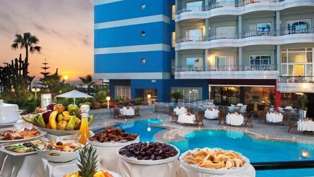Un buffet installé au bord de la piscine, proposant une variété de fruits et d’en-cas, entouré d’espaces de détente en plein air et d’un bâtiment d’hôtel doté de balcons.