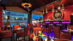 Un espace bar avec des chaises à revêtement rouge, un comptoir de bar orné de bouteilles et un grand miroir rond décoré d’un motif de roue de navire.