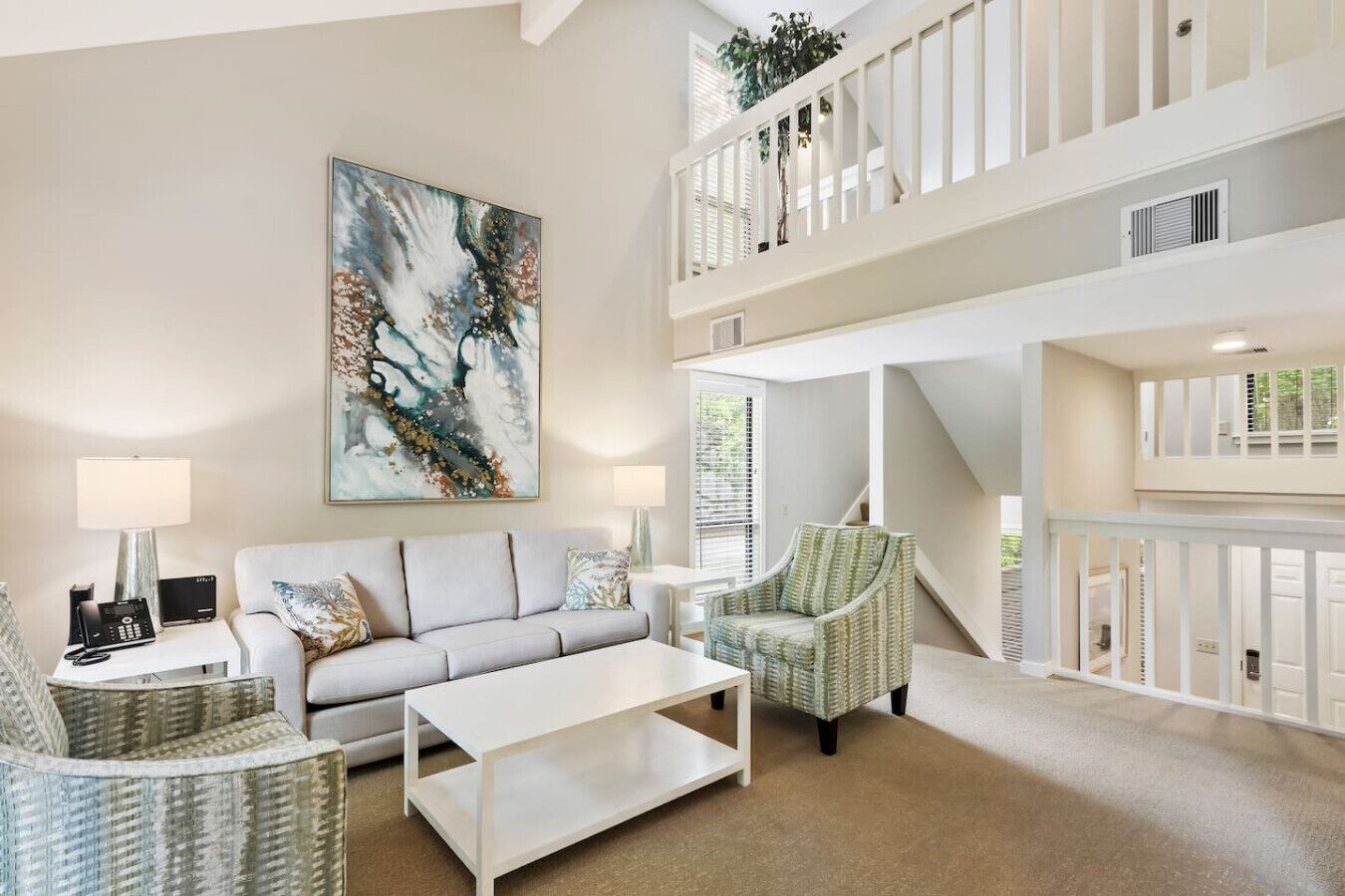 Un vaste espace de vie comprenant un canapé, des fauteuils, une table basse et un grand tableau abstrait. Un escalier mène à un niveau supérieur doté d’un balcon.