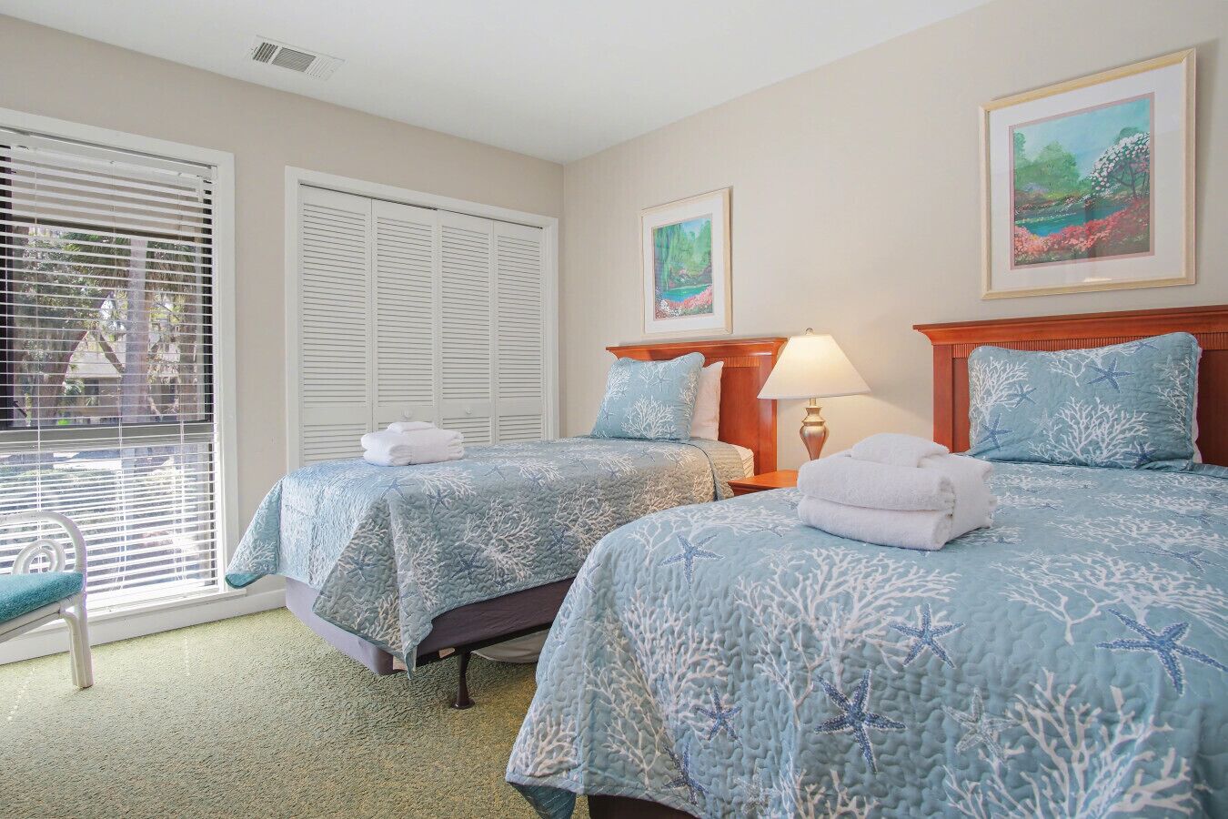 A hotel room with two beds, a window with blinds, a chair, and a painting on the wall.
