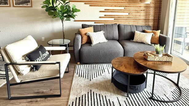 A modern living room with a grey sofa, a white armchair, and a round coffee table. There is a potted plant and a wall-mounted lamp.