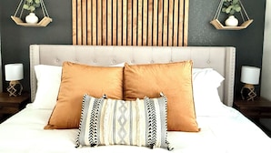 A bed with two large pillows and a smaller decorative pillow, flanked by two bedside tables with lamps, against a wall with vertical wooden slats and hanging plant shelves.