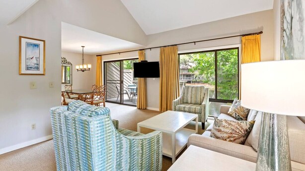 A spacious living room with a large window, a flat-screen TV, and a dining area with chairs and a table. - 577 - Fairway Views, Mins 2 Beach, Sleeps 6 (Hilton Head Island)
