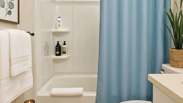 A bathroom with a shower, toilet, and a sink area. There are toiletries on a shelf, a candle on a stool, and a plant on the counter.