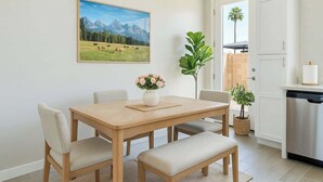 Een eetgedeelte met een houten tafel, vier stoelen, een bloevas en een schilderij van een berglandschap aan de muur.