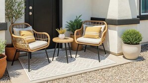 Een moderne buitenzitplaats met rieten stoelen, een kleine tafel, een zwarte deur en een plant in pot.