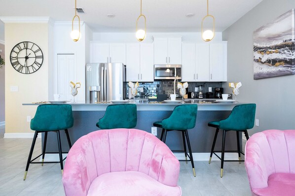 A modern kitchen with white cabinets, a stainless steel refrigerator, and a bar area with teal chairs. - Magical Stay Mickey and Harry Potter Rooms (Kissimmee)
