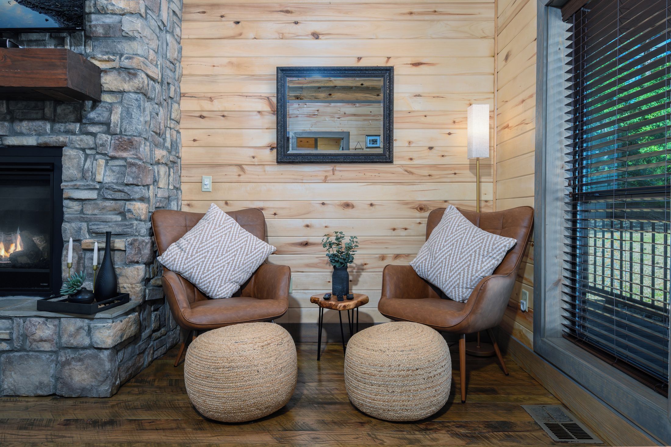 A cozy room with a stone fireplace, two leather armchairs with patterned cushions, a small wooden table with a vase, and a large window with blinds.