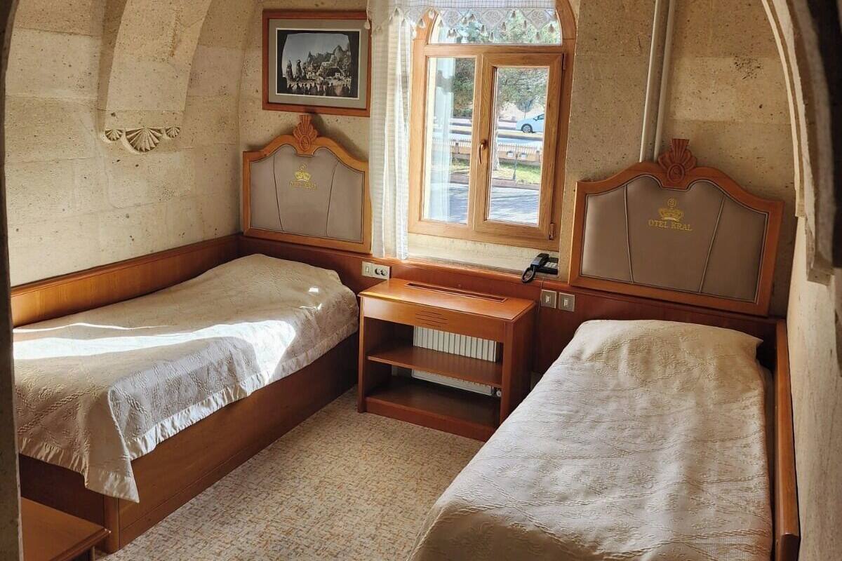 Ein Zimmer mit zwei Holzbetten, einem kleinen Tisch, einem Fenster mit Vorhängen und einem gerahmten Bild an der Wand.