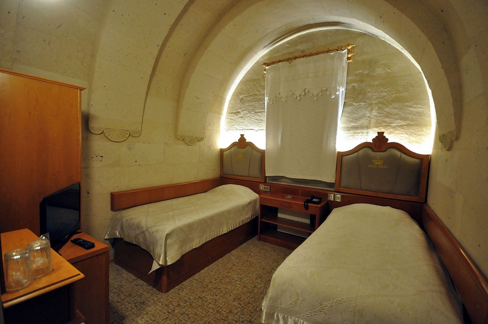 Une petite pièce agréable, dotée de deux lits simples, d’un bureau en bois, d’une chaise et d’une grande fenêtre en arc de cercle agrémentée d’un rideau.