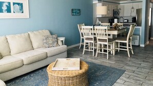 A living room with a sofa, a dining table with chairs, a kitchen area, and a blue wall with artwork. - SPECIAL PRICES FOR SEPT!  Beachfront Retreat! Monthly specials you can't believe (Panama City Beach)
