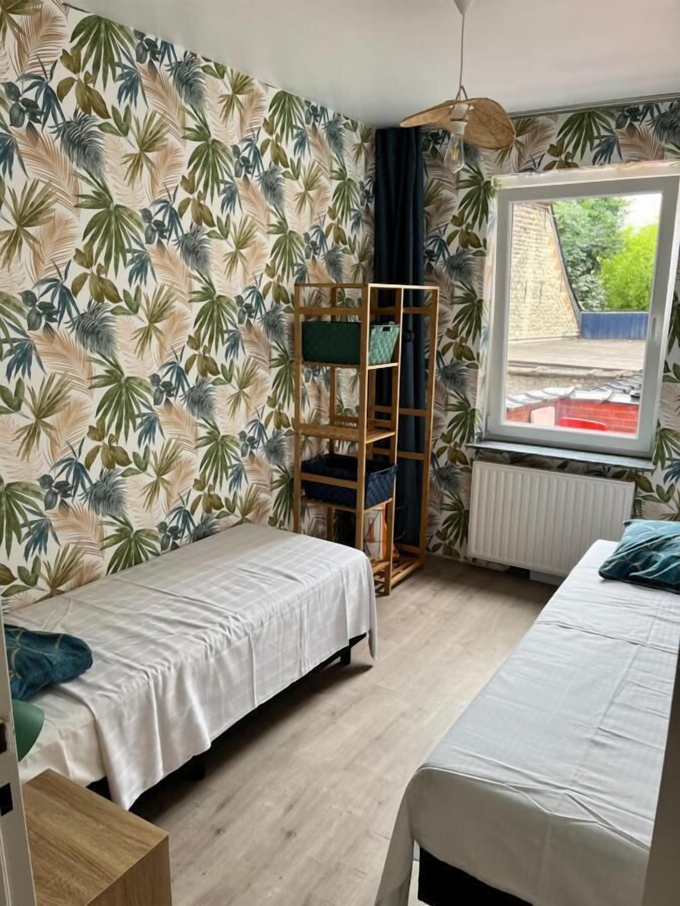 A room with two beds, a wooden shelf unit, a window with a view of the outside, and a tropical leaf-patterned wallpaper.