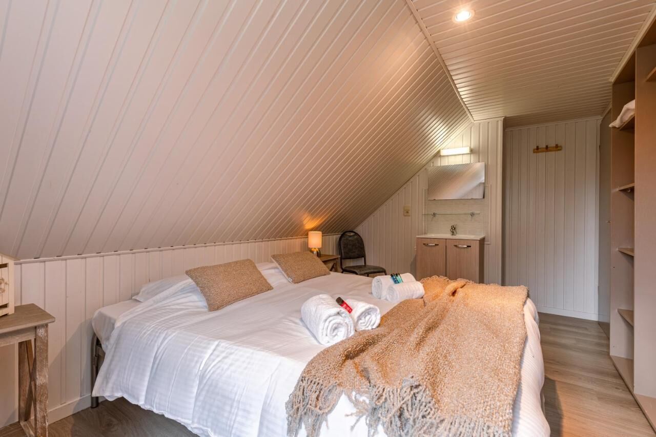 Une chambre avec un plafond en pente, un lit recouvert de draps blancs, une table de chevet en bois, une chaise, une petite table et un miroir accroché au mur.