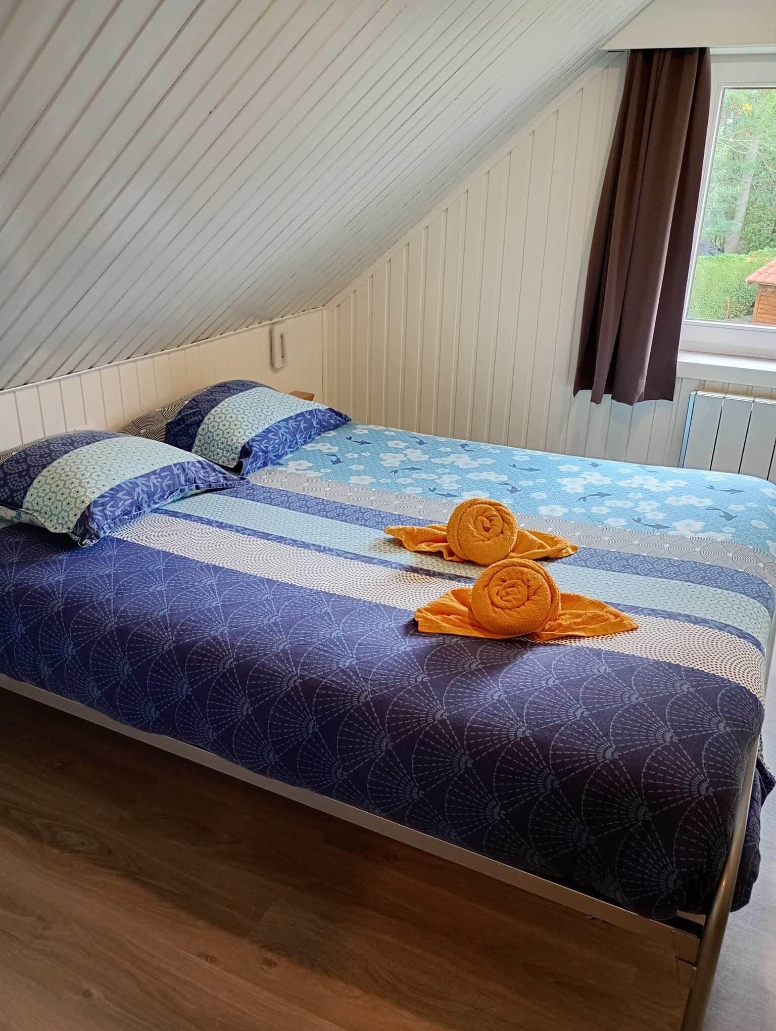 A single bed with a patterned bedspread, pillows, and two rolled towels.