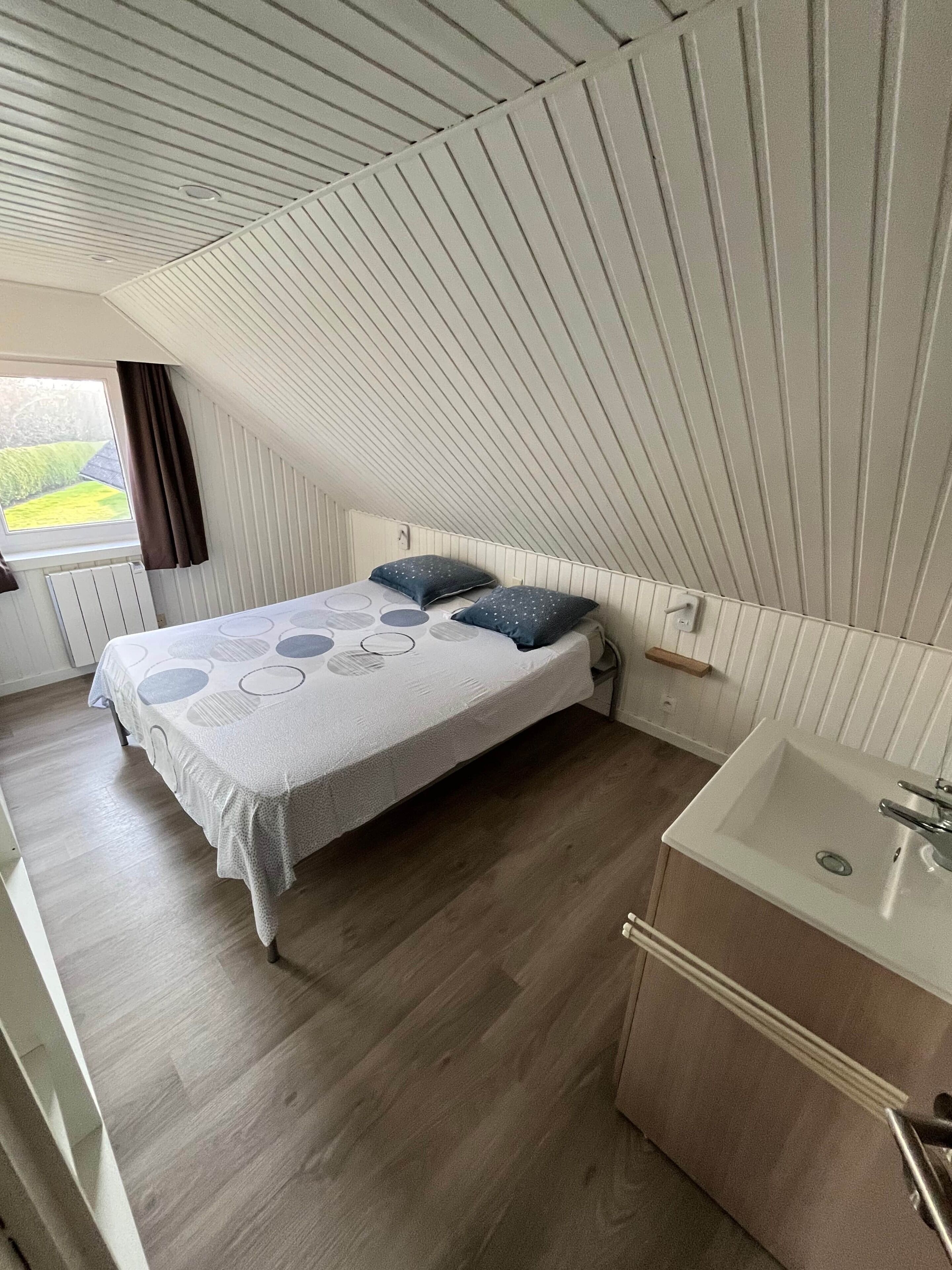 Ein Schlafzimmer mit Bett, Nachttisch, einem Fenster mit Vorhängen, Heizkörper und einem Badezimmer mit Waschbecken.
