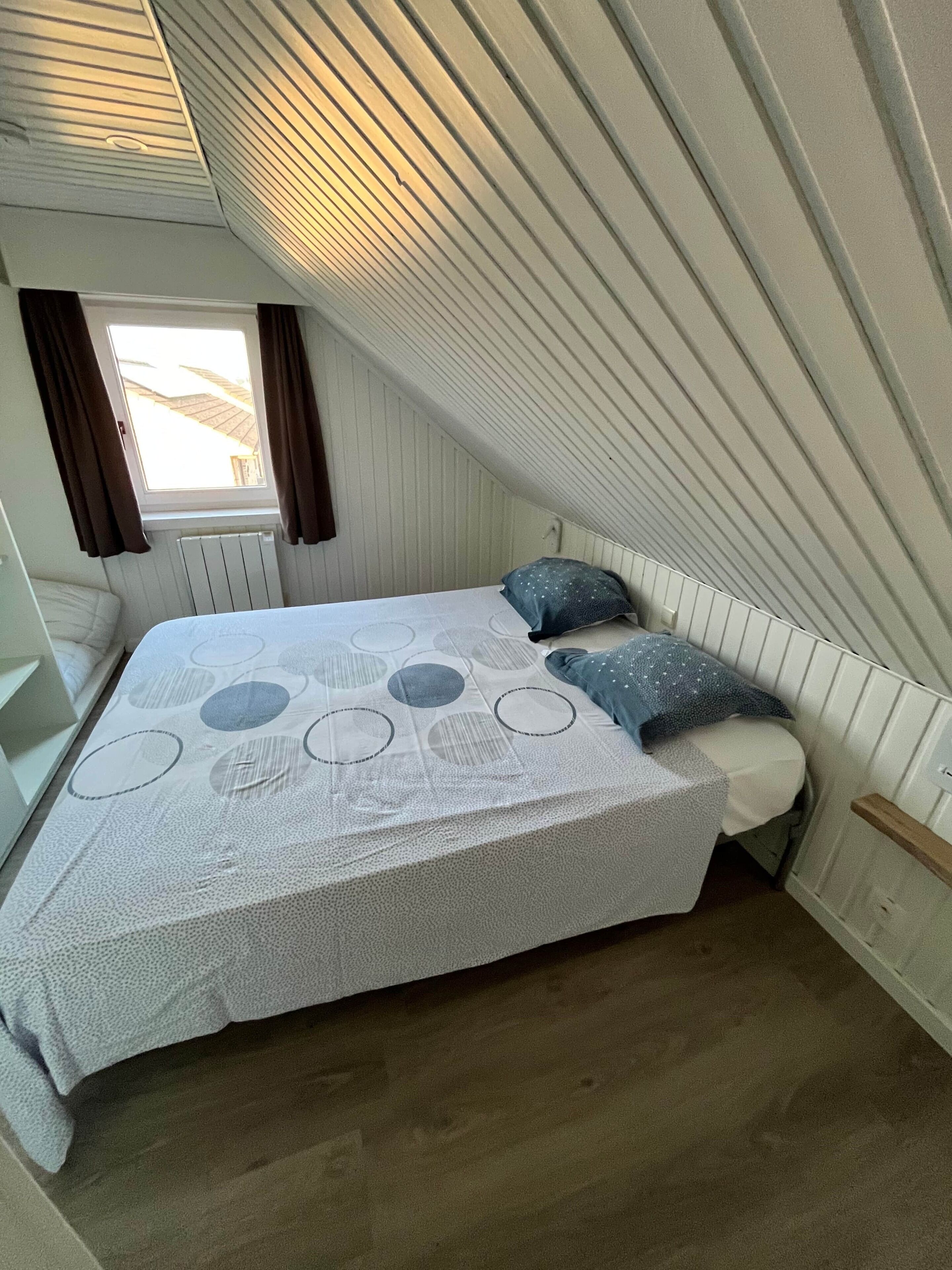 Een slaapkamer met een schuine kap, een bed met een bedspreider met patroon, twee kussens, een raam met gordijnen en een radiator.