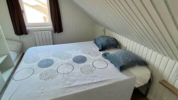 Een slaapkamer met een schuine kap, een bed met een bedspreider met patroon, twee kussens, een raam met gordijnen en een radiator.