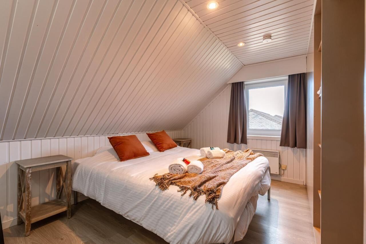Un dormitorio con techo inclinado, una cama con ropa de cama blanca, dos almohadas naranjas, una mesita de noche y una ventana con cortinas.