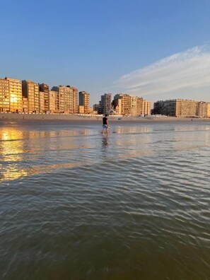 Una playa con una persona de pie en el agua, edificios al fondo y un cielo despejado.