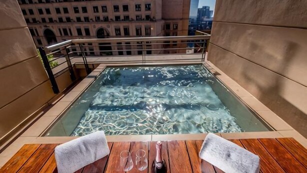 A rooftop hot tub with a wooden deck, a table with a towel and a bottle, and a cityscape view. - Raphael Suites (Sandton)