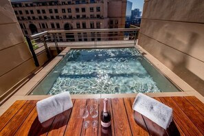 A rooftop hot tub with a wooden deck, a table with a towel and a bottle, and a cityscape view. - Raphael Suites (Sandton)