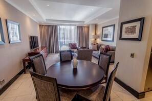 A hotel room with a round dining table, chairs, a sofa, a TV, and a window with curtains. - Raphael Suites (Sandton)