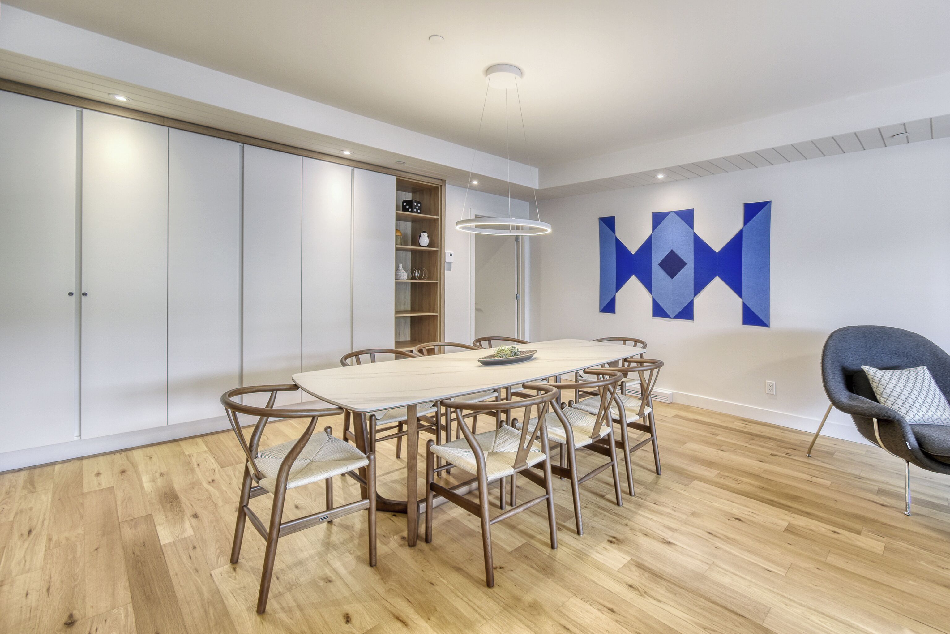 A modern dining area with a wooden table and chairs, a built-in bookshelf, and a geometric wall art piece.