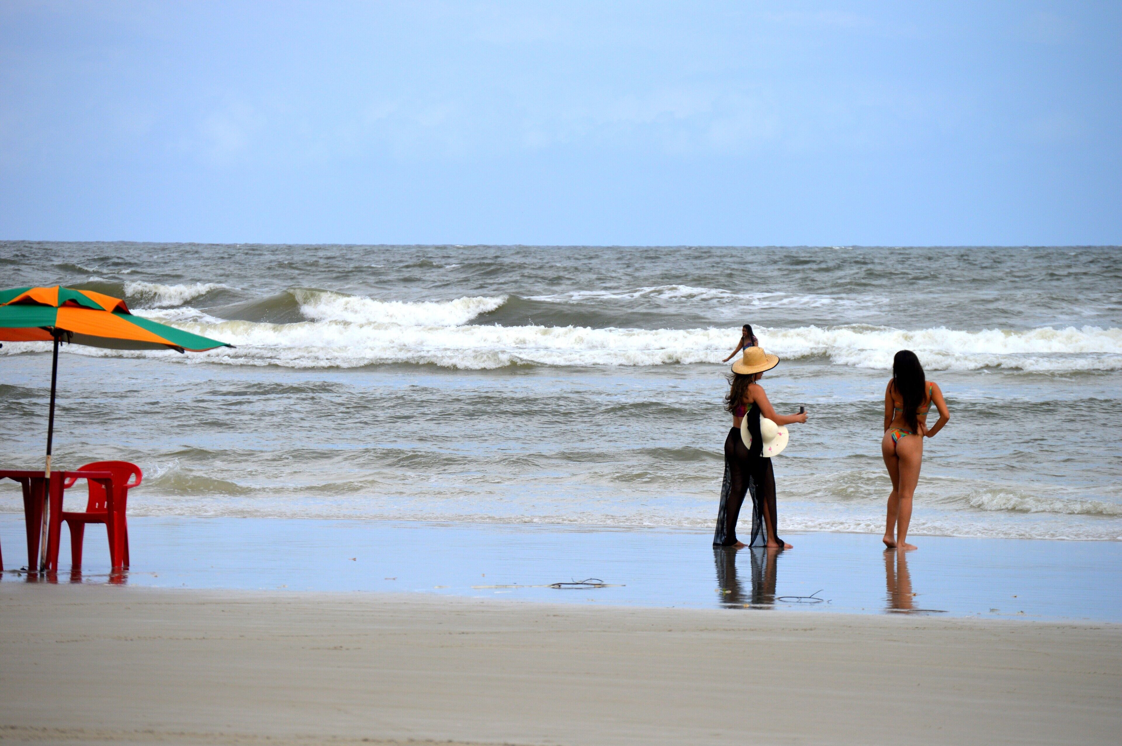 Une plage avec une chaise rouge, un parasol coloré et une femme portant un chapeau, qui tient un objet blanc.