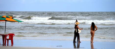 Une plage avec une chaise rouge, un parasol coloré et une femme portant un chapeau, qui tient un objet blanc.