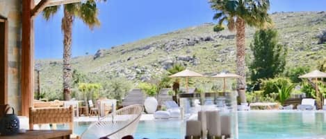 A poolside area with lounge chairs, a dining table with fruit, and a view of a mountainous landscape.