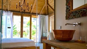 A room with a bed, a large wooden table, a rustic sink, and a view of greenery outside.