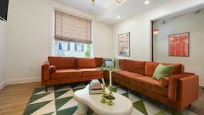 Un salon avec deux canapés orange, une table basse blanche agrémentée d’objets décoratifs, un tapis à motifs et une fenêtre avec des stores.