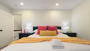 Un lit bien fait, recouvert d’une couverture jaune, avec deux oreillers rouges et une couette blanche. Il y a deux tables de chevet, chacune surmontée d’une lampe et d’un petit objet décoratif.