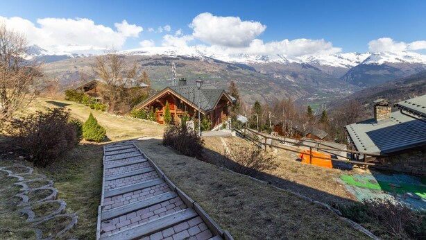 A wooden cabin with a sloped roof, surrounded by a stone wall and a paved pathway.