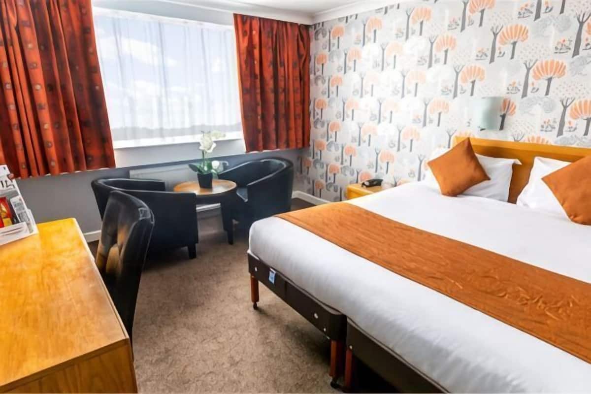 A hotel room with a bed, a desk, two chairs, a small table with a plant, and a window with curtains.