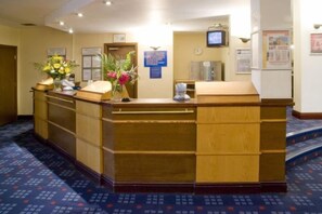 A reception desk with a wooden finish, a vase with flowers, and a clock on the wall. - Airport Hotel Manchester (Wythenshawe)