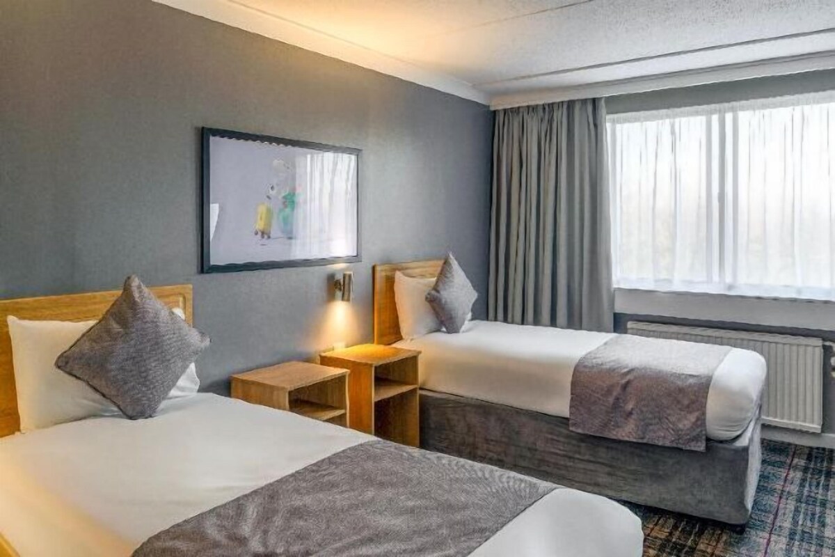 A hotel room with two beds, a small table, a wall-mounted artwork, and a window with curtains.