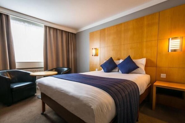 A hotel room with a large bed, two bedside tables, a chair, a small round table, and a window with curtains.