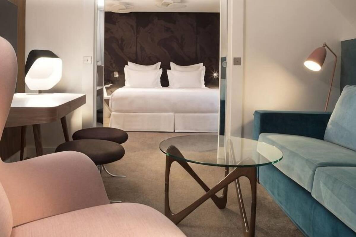A modern hotel room with a glass table, a blue sofa, a bed with white bedding, and a desk with a lamp.