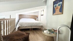 Un dormitorio pequeño y acogedor con una cama individual, piso de madera, un sillón de cuero marrón, una mesita con revistas y una silla blanca.