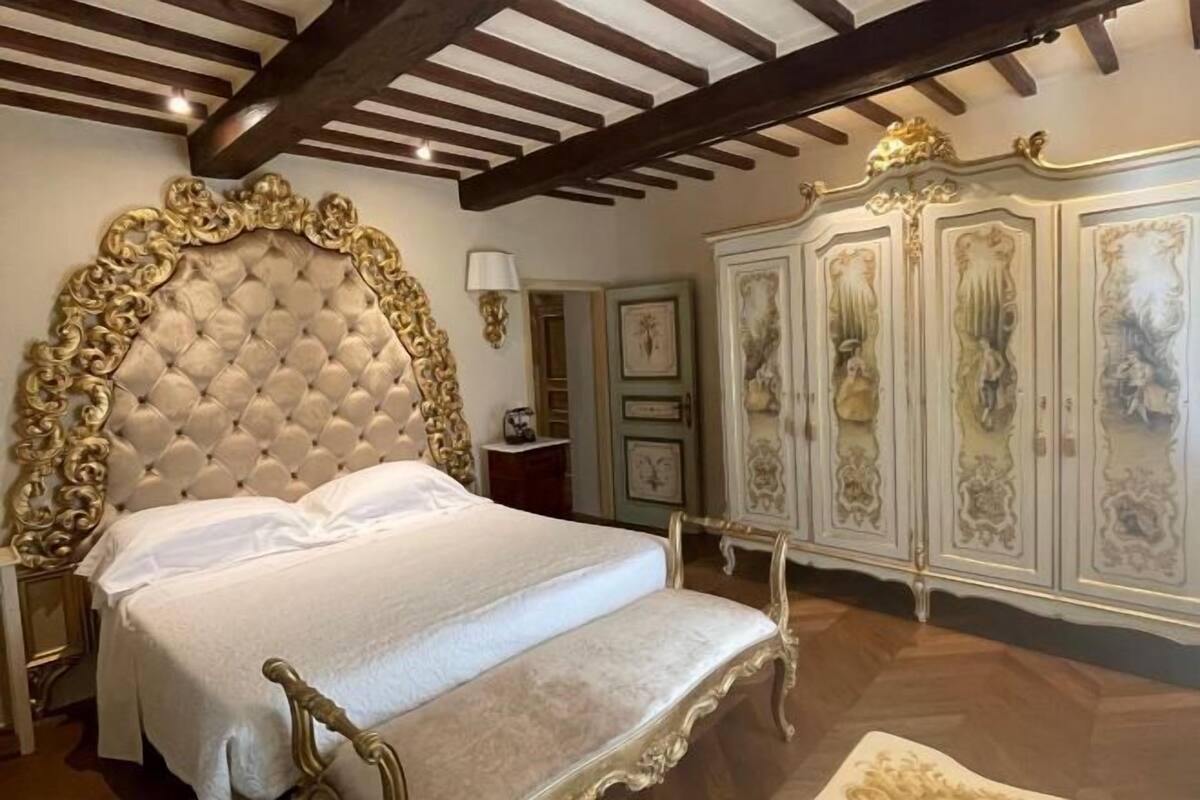 A bedroom with a large tufted headboard, a bed with white linens, a wooden floor, and ornate wall decorations.