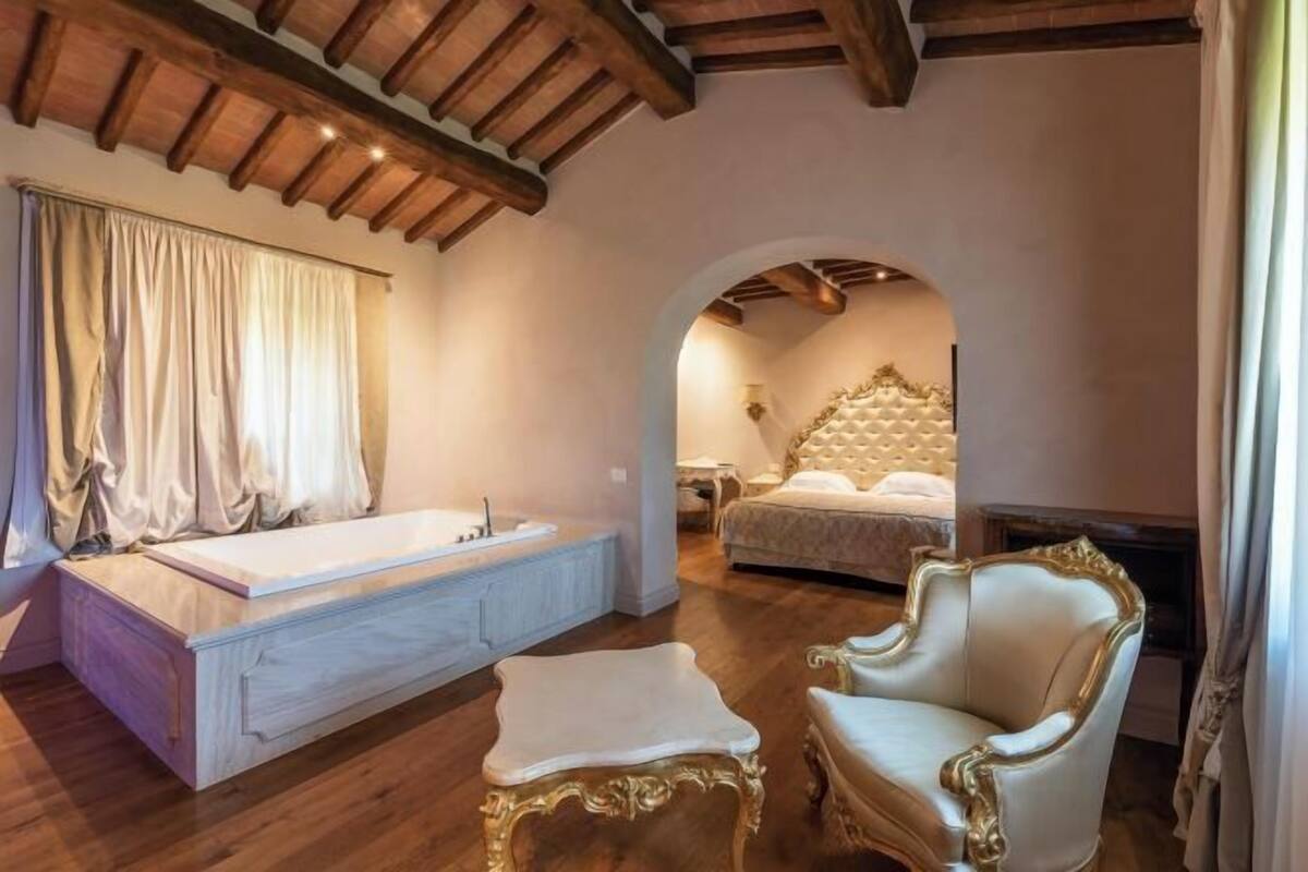 A bedroom with a large bed, a sitting area with a chair, a bathtub, and a wooden ceiling.