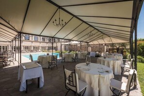 A covered outdoor dining area with tables and chairs, a pool in the background, and a building with a light-colored facade. - Relais la Corte dei Papi (Cortona)