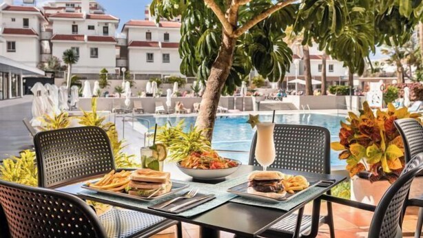 Un área de comedor al aire libre con vista a la piscina, con una mesa dispuesta con comida, sillas y una planta tropical.