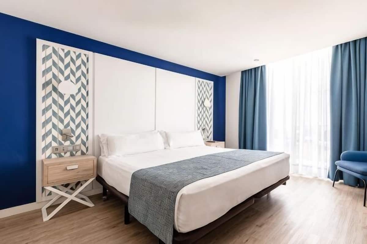 Ein Schlafzimmer mit einem Bett, einem Nachttisch, einem Sessel und einem Fenster mit Vorhängen.
